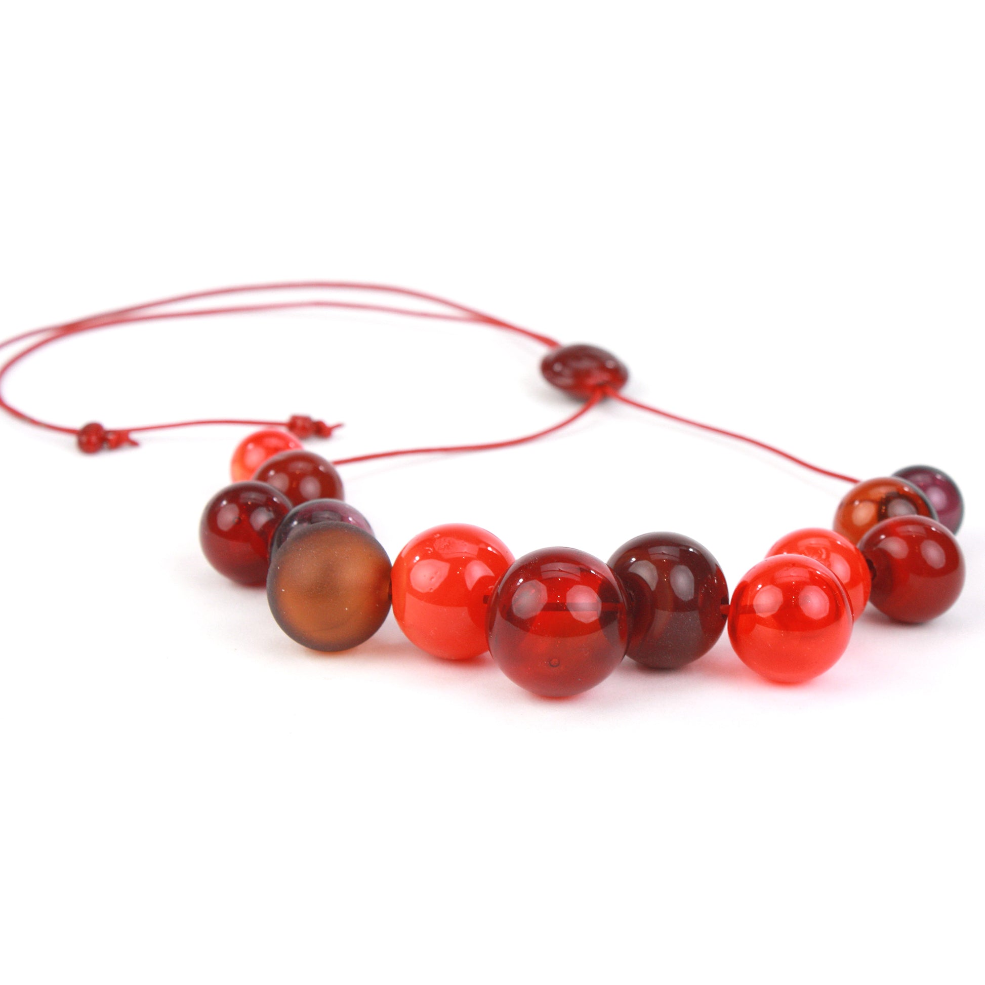 Offset Bolla Necklace featuring hand-blown glass beads in red, amber, and burgundy hues strung on red leather cord with a handcrafted adjustable slider bead.