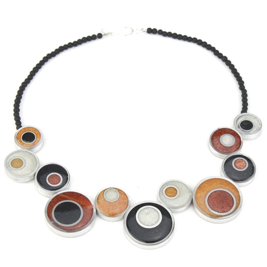 A handcrafted offset necklace featuring eco-resin and salvaged aluminum beads in black, white, copper, and gold tones, accented with glass beads and a silver clasp.
