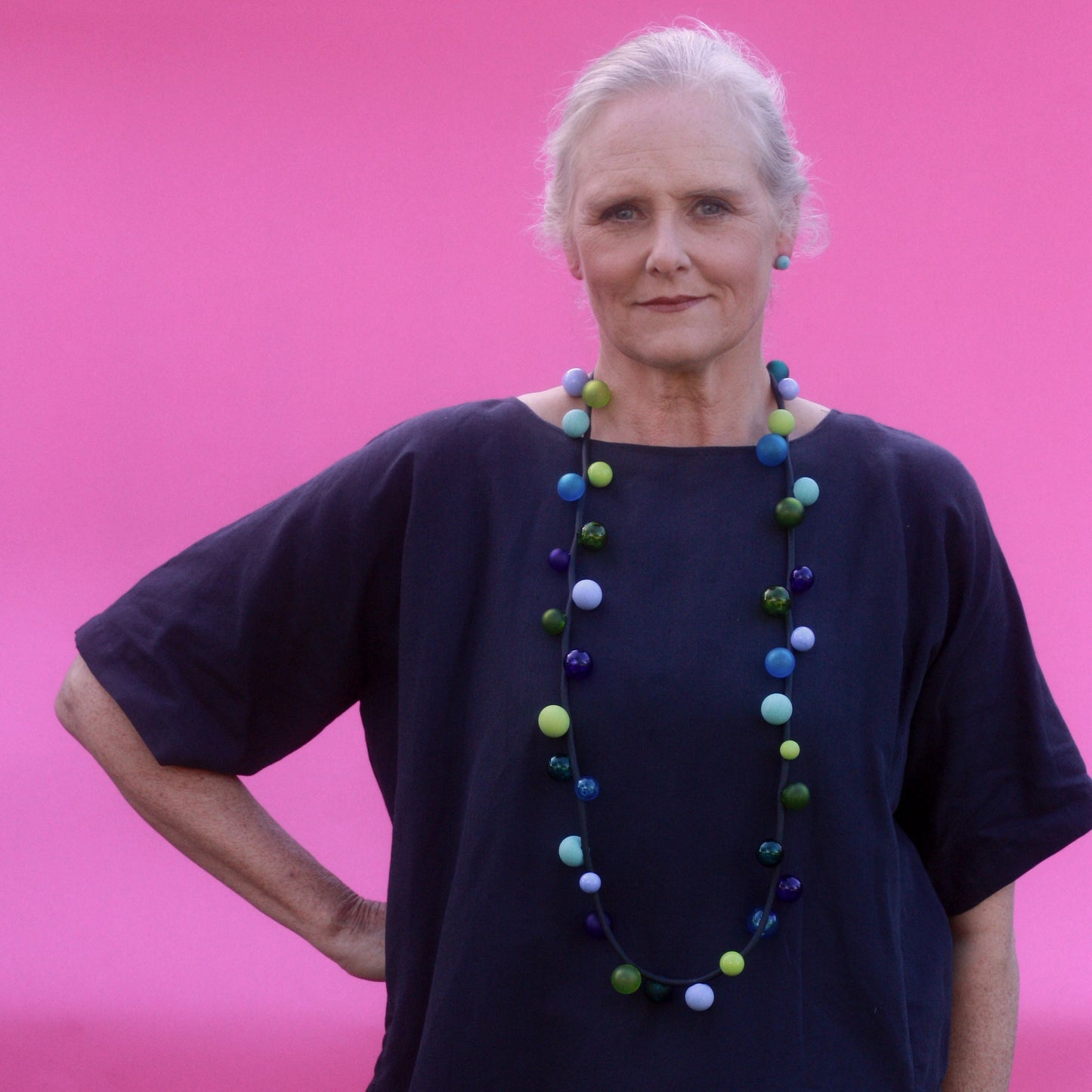 The "Bolla" necklace featuring hand-blown glass beads in shades of blue, green, and teal, strung on a steel cable threaded through a rubber cord, worn long or doubled.