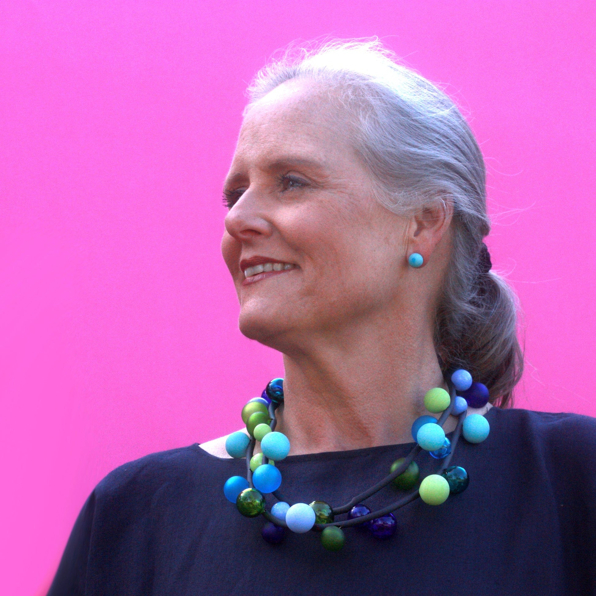 The "Bolla" necklace featuring hand-blown glass beads in shades of blue, green, and teal, strung on a steel cable threaded through a rubber cord, worn long or doubled.