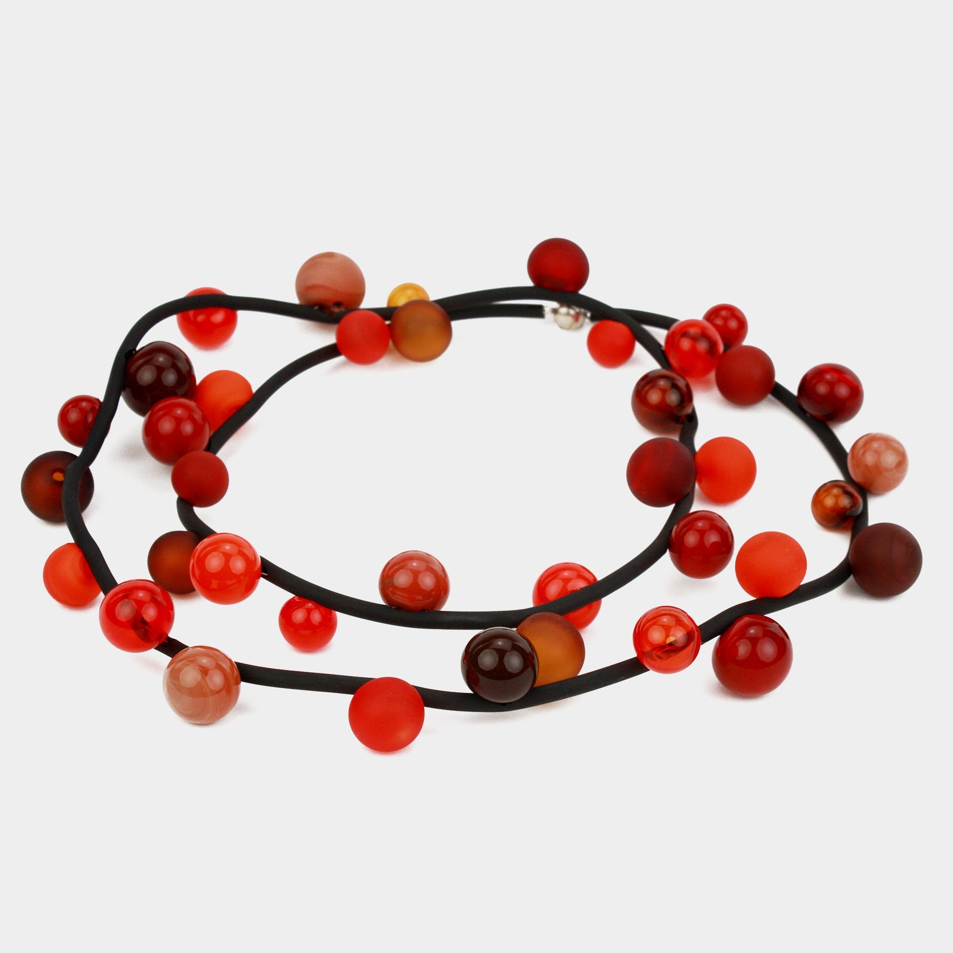 "Bolla" necklace with hand-blown glass beads in red, orange, and amber tones, strung on a steel cable within a rubber cord, shown in a long style.